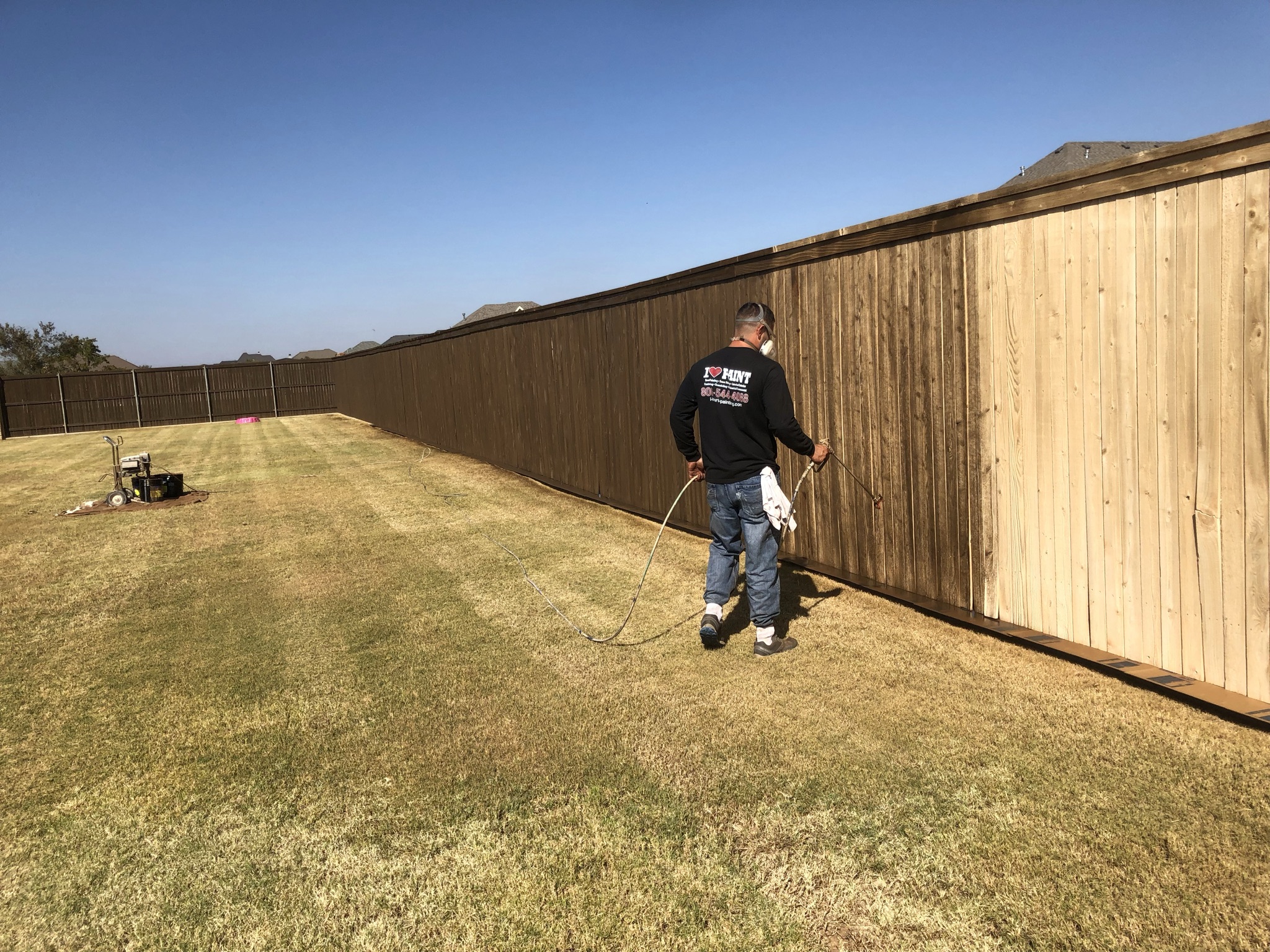 Worker spray staining a wood fence showing before and after halves